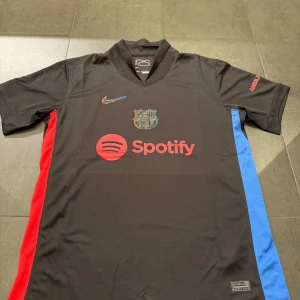FC Barcelona svart fotbollströja med Spotify-tryck - Snygg svart FC Barcelona fotbollströja från Nike med klubbmärke på bröstet, röd och blå rand på sidorna samt Spotify-logga framtill. UNHCR-tryck på ryggen. Perfekt för dig som älskar Barca och vill sticka ut på planen.