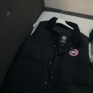 Svart dunväst från Canada Goose - Säljer en svart dunväst från Canada Goose med klassisk logotyp på bröstet. Västen har knappar och hög krage, perfekt för lager-på-lager under kyliga dagar.