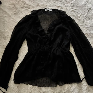 Svart blus med volang från NA-KD - Säljer en svart blus från NA-KD i storlek 34. Blusen har långa ärmar, v-ringning och fina volangdetaljer längs halsringningen och framtill. Tunt, lätt material som ger en elegant känsla.💕