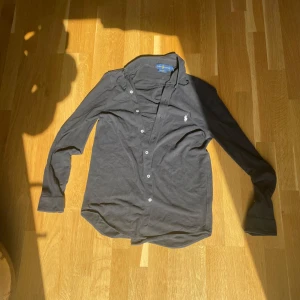 Ralph Lauren skjorta - Säljer en stilren svart långärmad skjorta från Ralph Lauren. Skjortan har en klassisk krage och knappar framtill. Den är dekorerad med Ralph Laurens ikoniska logga broderad i vitt på bröstet. Perfekt för en elegant look nu till våren.  Hör av dig vid minsta fundering😎