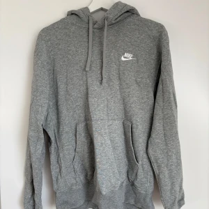 Grå hoodie från Nike - Grå hoodie från Nike med vit logga på bröstet. Tröjan har huva med dragsko och en stor magficka framtill. Perfekt för en avslappnad stil och enkel att matcha med det mesta.
