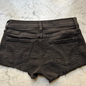 Svarta jeansshorts från Hollister - Svarta jeansshorts från Hollister med råa, fransiga kanter och klassisk femficksdesign. Shortsen har låg midja och knappgylf. Perfekta för varma dagar och en avslappnad stil.