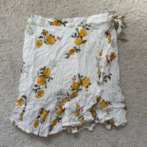 Vit blommig omlottkjol från Lindex - Säljer en söt vit omlottkjol från Lindex med gult blommönster och volangdetaljer. Kjolen har knytning i sidan och är perfekt för varma dagar. Tunn och luftig känsla.
