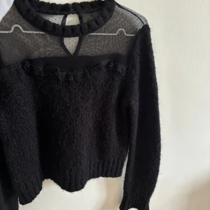 Svart stickad tröja med mesh och volang - Säljer en svart stickad tröja med transparent mesh upptill och volangdetalj längs bröstet. Tröjan har långa ärmar. Perfekt för dig som vill ha något unikt och stilrent i garderoben.