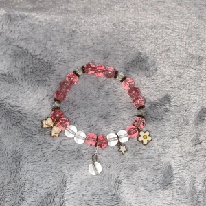 Rosa och vit pärlarmband med berlocker - Sött armband med rosa och vita pärlor, dekorerat med berlocker i form av rosett, stjärna, blomma och en rund genomskinlig pärla. Perfekt accessoar för att ge en lekfull touch till din stil.