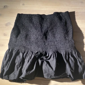 Svart volangkjol från H&M - Supersöt svart kjol från H&M med smock upptill och volang nertill. Kjolen är kort och har en skön passform som sitter snyggt i midjan. Perfekt att styla med en enkel topp eller hoodie.
