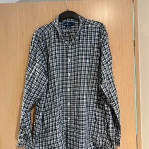 Rutig skjorta från Ralph Lauren - Säljer en klassisk rutig skjorta från Ralph Lauren i blått, vitt och grönt. Skjortan har långa ärmar, knappar framtill och en broderad logga på bröstet. Perfekt för dig som gillar en tidlös och avslappnad stil.