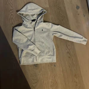 Ljusgrå hoodie från Polo Ralph Lauren - Säljer en ljusgrå hoodie från Polo Ralph Lauren med dragkedja och huva. Tröjan har två fickor framtill och den klassiska lilla loggan broderad på bröstet. Perfekt för en avslappnad stil.