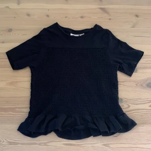 Svart topp med volang från Gina Tricot - Säljer en mörkblå topp från Gina Tricot med korta ärmar och volangdetalj längst ner. Toppen har smockad struktur på fram- och baksidan vilket ger en snygg och trendig look. Perfekt att styla med jeans eller kjol! 