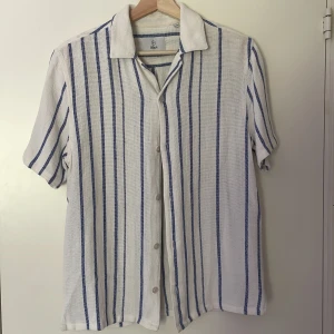 Skjorta - Kortärmad skjorta från Bläck i vit med blå vertikala ränder. Skjortan har klassisk krage, knäppning framtill och är tillverkad i ett luftigt material som passar perfekt till varma dagar. Perfekt till stranden eller utekvällar. Nypris 600kr.
