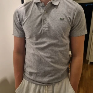 Grå pikétröja från Lacoste - Grå pikétröja från Lacoste med den ikoniska krokodilloggan på bröstet. Tröjan har korta ärmar och en stilren krage med knappar.