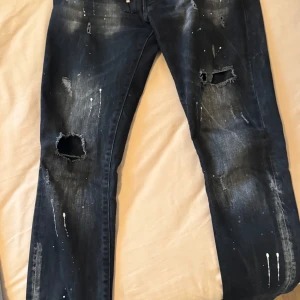 Svarta slitna jeans från Dsquared2 - Säljer ett par svarta jeans från Dsquared2 med slitningar och hål på benen samt vita färgstänk. Jeansen har en grov dragkedja och klassisk femficksmodell. Snygg streetstil och coola detaljer.