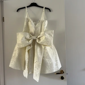 Vit kortklänning med stor rosett examen/student klänning från CrazyDizzy - Supersöt vit kortklänning från CrazyDizzy med axelband och en stor dekorativ rosett framtill. Klänningen har ett subtilt blommigt mönster i tyget och är tillverkad i ett glansigt material som ger en lyxig känsla. Perfekt för speciella tillfällen! Rosetten går att ta av.