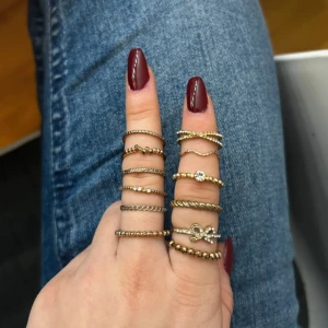 Set med guldfärgade ringar med stenar och pärldetaljer - Säljer ett set med flera tunna guldfärgade ringar i olika designer. Vissa ringar har små glittriga stenar och andra har pärlformade detaljer. Perfekt för att mixa och matcha på flera fingrar.