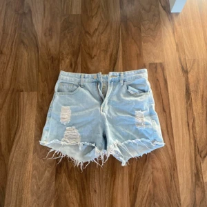 Ljusblå jeansshorts med fransar - Säljer ett par ljusblå jeansshorts med slitna detaljer och fransig kant. Klassisk femficksmodell med bälteshällor och dragkedja. Perfekta för sommaren och ger en avslappnad vibe.