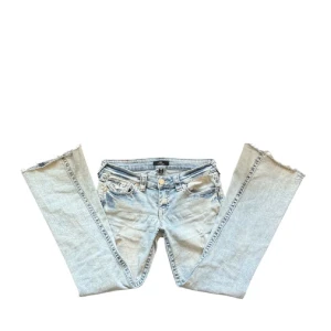 Ljusblå jeansbyxor från True Religion med slitningar  - Säljer ett par ljusblå jeans från True Religion med slitna detaljer och klassisk broderad bakficka. Byxorna har låg midja, med bootcut modell. Jeansen har två snygga hål vid knäna. Säljer då de är för korta för mig som är 168. Kan skicka fler bilder med dem på🙏🤍Storleken är w27