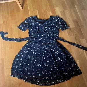 Mörkblå blommig klänning med knappar och knytskärp - Supersöt mörkblå klänning med små vita blommor, knappar framtill och knytskärp i midjan. Klänningen har korta ärmar och lätt v-ringning. Perfekt för vår och sommar!