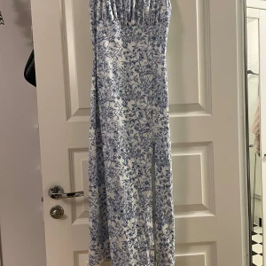 Vit och blå blommig långklänning - Säljer en romantisk långklänning i vitt med blått blommönster. Klänningen är ärmlös och har en lätt, luftig känsla – perfekt för varma dagar. Passformen är normal och tyget känns mjukt mot huden.