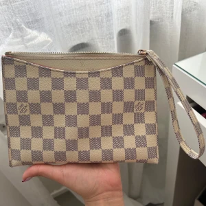 Vit patch väska - Säljer en snygg kuvertväska i beige och grått rutigt mönster. Väskan har dragkedja upptill, avtagbar handledsrem och röd insida. Perfekt för att bära det viktigaste med stil.