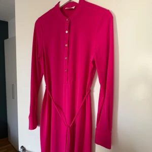 Ny Calvin Klein fuchsia klänning, storlek 4 - *Elegant fuchsiafärgad klänning från Calvin Klein. *Klänningen har långa ärmar med knappar och en smickrande knytdetalj i midjan.  *Den har en stilren design med knappar framtill. *Aldrig använd, endast tvättad. *Storlek 4, passar till Xs *Ordinarie pris runt 2000 kr *Djurfritt och rökfritt hem.