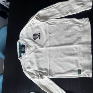Vit stickad tröja från Morris - Vit stickad tröja från Morris med broderat emblem på bröstet. Tröjan har ribbade muddar vid ärmslut och nederkant. Klassisk och stilren design, perfekt för en preppy look. Enda defekten är hålet i armhålan som inte syns vid användning.