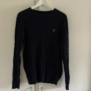 Mörkblå kabelstickad tröja från GANT - Säljer en klassisk mörkblå kabelstickad tröja från GANT i storlek M. Tröjan har rund halsringning, lång ärm och diskret logga broderad på bröstet. Perfekt för lager-på-lager och stilren look.