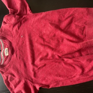 Röd stickad tröja från Hilfiger Denim - Säljer en röd stickad tröja från Hilfiger Denim. Tröjan har rund halsringning och en liten broderad logga på bröstet. Perfekt för dig som gillar klassisk och stilren design.