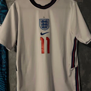 England fotbollströja med nummer 11 - Säljer en vit England fotbollströja från Nike med nummer 11 på bröstet och landslagets emblem. Tröjan har korta ärmar och detaljer i blått och rött längs sidorna och kragen. Perfekt för fotbollsfans!