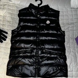 Svart dunväst från Moncler - Säljer en svart, glansig dunväst från Moncler med hög krage och dragkedja framtill. Västen har två fickor med dragkedja och tryckknappar med Moncler-logga. Klassisk Moncler-logotyp på bröstet.