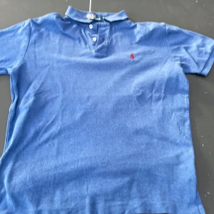 Blå pikétröja från Polo Ralph Lauren - Säljer en klassisk blå pikétröja från Polo Ralph Lauren med korta ärmar och krage. Tröjan har två knappar vid halsen och en röd broderad logga på bröstet. Perfekt för en stilren och avslappnad look.