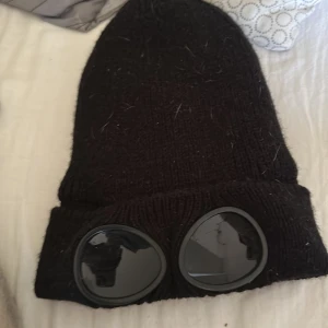 Svart stickad mössa med glasögondetalj - Svart stickad mössa med uppvikt kant och två runda glasögonliknande detaljer framtill. Perfekt accessoar för en unik och cool stil. Använd två gånger förut. 