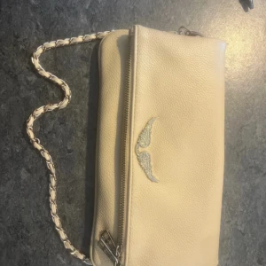 Zadig Väska - Säljer en beige axelväska med kedjedetalj i axelbandet och ett litet broderat motiv framtill. Väskan har dragkedja och magnetknappar samt ett fack på baksidan. Perfekt storlek för det viktigaste.