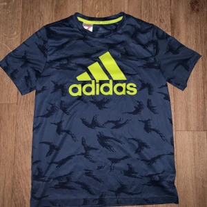Mörkblå Adidas t-shirt med neongult tryck - Snygg mörkblå t-shirt från Adidas med neongult logotryck på bröstet. T-shirten har ett diskret mönster och är tillverkad i ett syntetmaterial som andas. Perfekt för träning eller sportiga dagar.