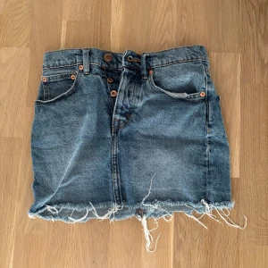Jeanskjol med fransig kant - Säljer en klassisk blå jeanskjol med rå, fransig nederkant och fem fickor. Kjolen har knapp- och dragkedjestängning framtill och orangea sömmar. Perfekt för en avslappnad stil.