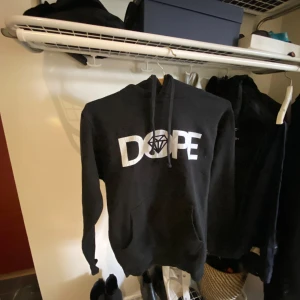 Svart hoodie med DOPE-tryck - Svart hoodie med stort vitt DOPE-tryck och diamantmotiv på bröstet. Klassisk huva med dragsko och lång ärm. Perfekt för en avslappnad streetwear-look.