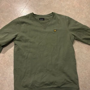 Olivgrön sweatshirt från Lyle & Scott - Säljer en olivgrön sweatshirt från Lyle & Scott Junior med klassisk rund hals och broderad logga på bröstet. Tröjan har långa ärmar och är tillverkad i mjuk bomull, perfekt för en avslappnad stil.
