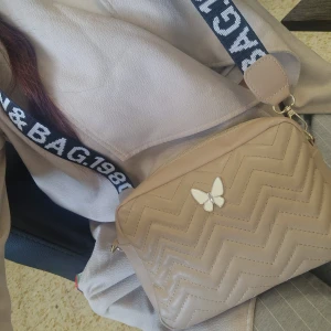 Beige axelväska med fjärilsdetalj - Säljer en beige axelväska med quiltat sicksackmönster och en söt fjärilsdetalj framtill. Väskan har en bred axelrem med text och dragkedja upptill. Perfekt för att bära det viktigaste med stil.