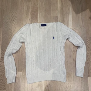 Vit kabelstickad tröja från Polo Ralph Lauren - Kabelstickad vit tröja från Polo Ralph Lauren med klassisk blå broderad logga på bröstet. Tröjan har rund halsringning och långa ärmar. Perfekt för lager-på-lager och en stilren look.