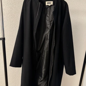 Svart lång jacka från Weekday - Säljer en svart lång jacka från Weekday med minimalistisk design och rak passform. Jackan har dragkedja framtill och är helsvart både in- och utvändigt. Perfekt för dig som gillar stilrena plagg.