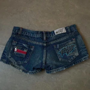 Blå low rise/waist jeansshorts med broderad bakficka - Säljer ett par blå jeansshorts. Baksidan har två fickor, varav en med röd dragkedja och den andra med broderad text och mönster. Klassisk femficksmodell och låg midja. Dyrt märke och mening är att den ska vara sliten vid lite små ställen framme och bak! Öppen för bud.🌷