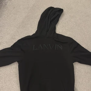 Svart hoodie från Lanvin - Säljer en svart hoodie från Lanvin med broderad logga på bröstet. Tröjan har huva med dragsko och en klassisk passform. Perfekt för dig som gillar stilrena och exklusiva plagg.