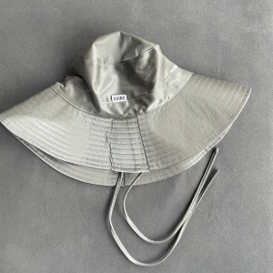 Grå regnhatt från Rains - Säljer en grå regnhatt från Rains med bred brätte och knytband under hakan. Hatten är tillverkad i vattentätt material och har en mjuk insida för extra komfort. Perfekt för regniga dagar.