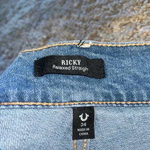True Religion Ricky Relaxed Straight jeans - Säljer ett par ljusblå True Religion Ricky Relaxed Straight jeans med klassiska kontrastsömmar och ikonisk bakficka med lock och knapp. Jeansen har normal passform och raka ben, perfekt för en avslappnad stil.