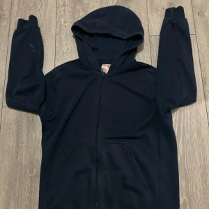 Mörkblå hoodie från Adidas Athletics - Mörkblå hoodie med dragkedja från Adidas Athletics. Stor vit Adidas-logga och text på ryggen. Tröjan har huva, långa ärmar och två fickor framtill. Perfekt för träning eller chill.