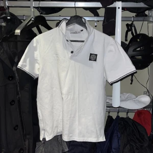 Vit t-shirt med krage från Stone Island - Vit t-shirt från Stone Island med korta ärmar och svart/vit krage. T-shirten har en svart patch med logga på bröstet och svarta detaljer vid ärmslut och krage. Perfekt för en stilren och sportig look.