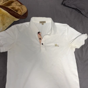 Vit piké t-shirt från Burberry - Säljer en klassisk vit piké t-shirt från Burberry med ikonisk rutdetalj vid knäppningen och broderad logga på bröstet. T-shirten har krage och korta ärmar, perfekt för en stilren look.