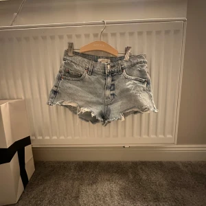 Ljusblå jeansshorts med fransar - Säljer ett par ljusblå jeansshorts med råa fransade kanter och klassisk femficksdesign. Shortsen har låg midja och slitna detaljer framtill för en trendig look. Perfekta för sommaren!