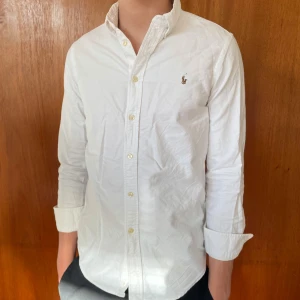 Vit skjorta från Ralph Lauren - Klassisk vit skjorta från Ralph Lauren med broderad logga på bröstet. Skjortan har lång ärm, knappar framtill och är tillverkad i bomull. Perfekt för en stilren och tidlös look. Modellen är ca 173 då skjortan är i storlek 16 men passar dig som har xs  