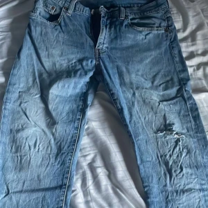 Blå jeansbyxor från Levi's - Klassiska blå jeansbyxor från Levi's med fem fickor och dragkedja. Byxorna har en rak passform och ett slitet hål på höger lår för en cool, avslappnad look. Perfekta till vardags för en tidlös stil.