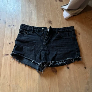 Svarta jeansshorts med fransar - Säljer ett par svarta jeansshorts med råa fransade kanter. Klassisk femficksmodell med knapp och dragkedja framtill. Perfekta för en avslappnad stil.